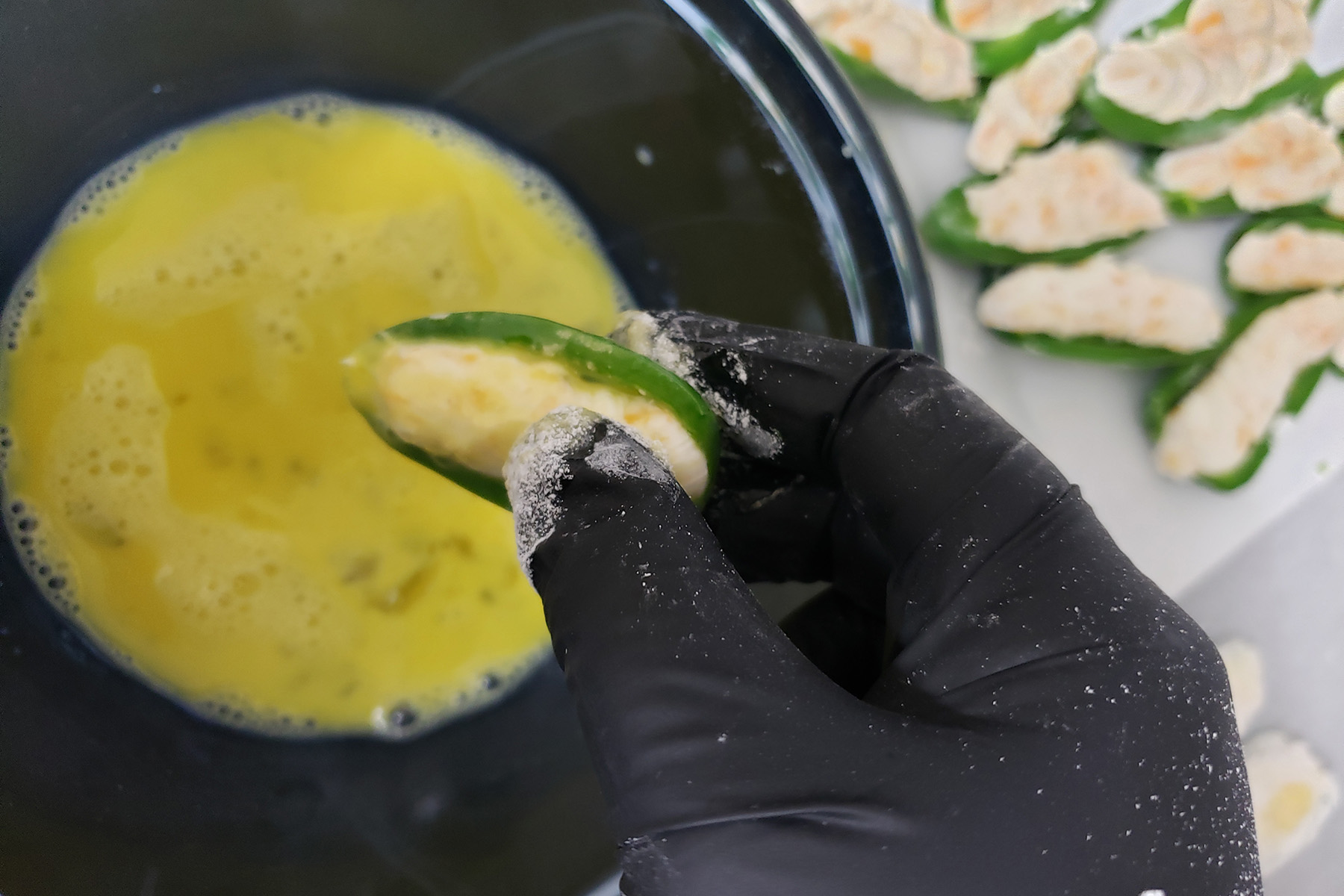 A gloved hand is shown dipping a filled jalapeno half into egg.