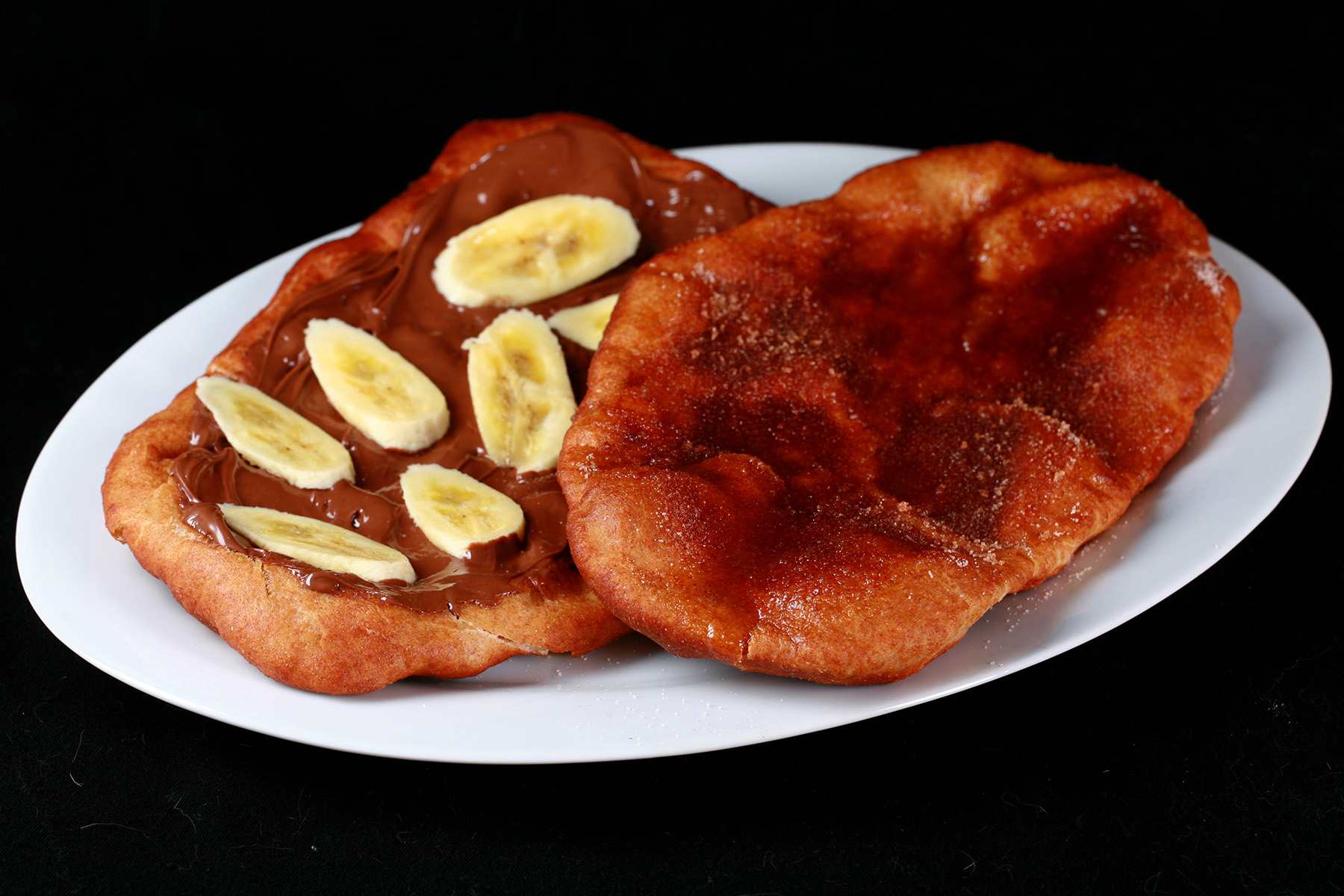 "Two gluten-free beaver tail pastries are arranged on a plate. Both are oblong pieces of fried dough. One is topped with cinnamon sugar, the other is spread with Nutella and topped with sliced banana.