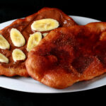 Two gluten-free beaver tail pastries are arranged on a plate. Both are oblong pieces of fried dough. One is topped with cinnamon sugar, the other is spread with Nutella and topped with sliced banana.