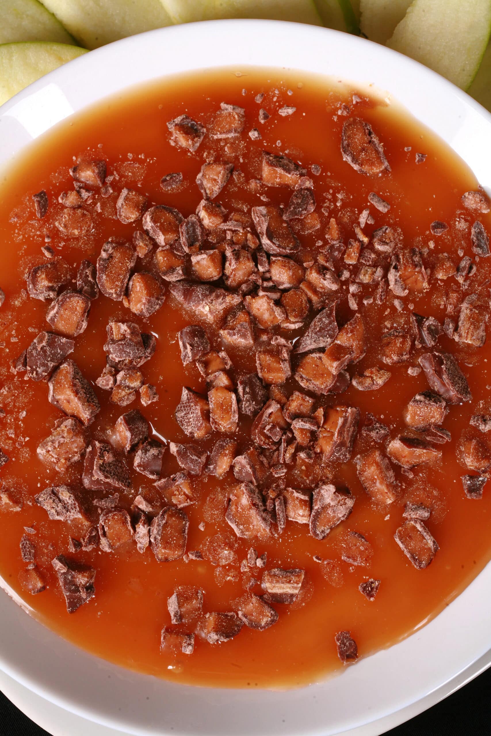 A bowl of cream cheese caramel apple dip, with caramel sauce and chopped Skor bars on top, and sliced granny smith apples behind it.