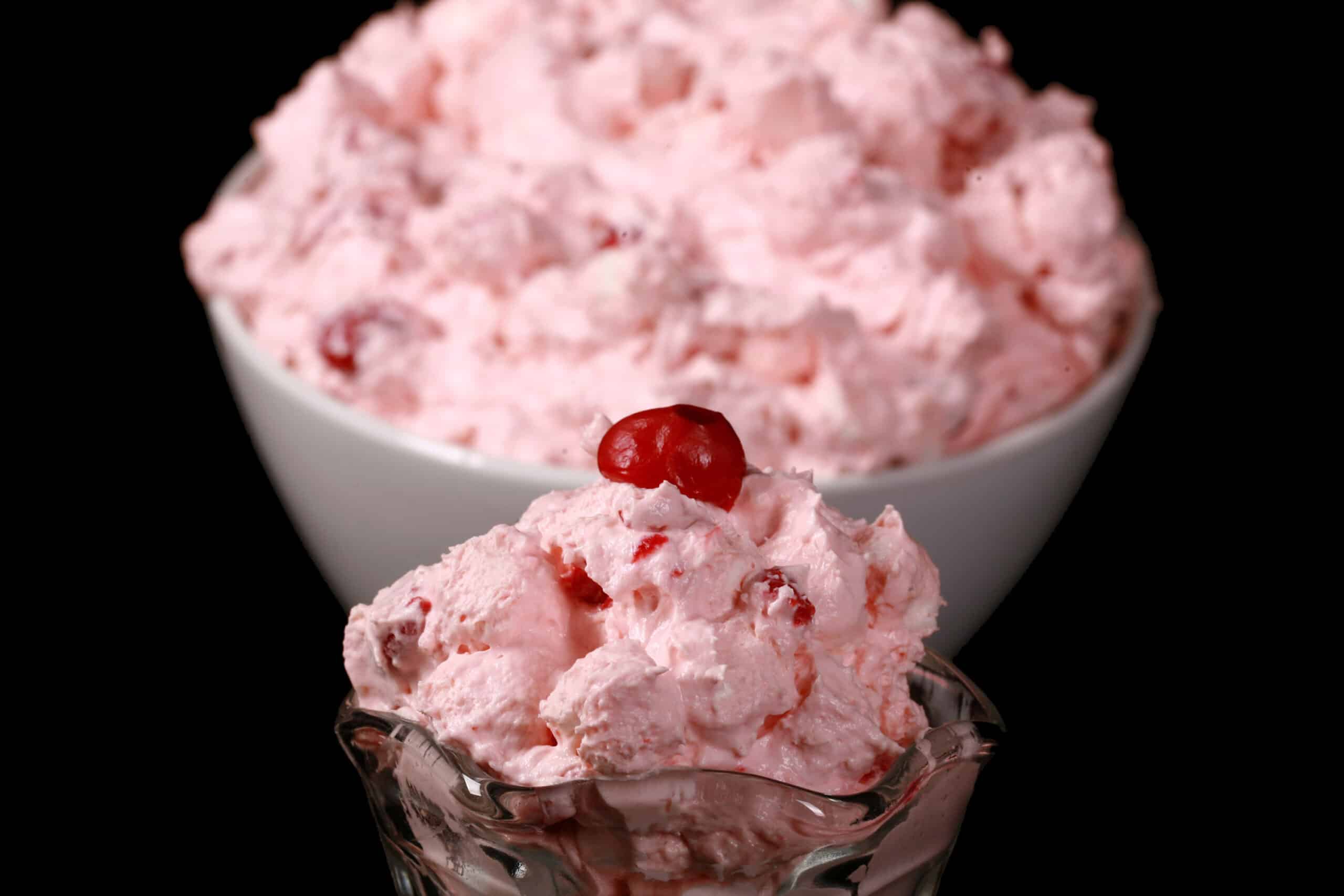 A serving glass of cherry fluff salad, in front of a bowl of it.