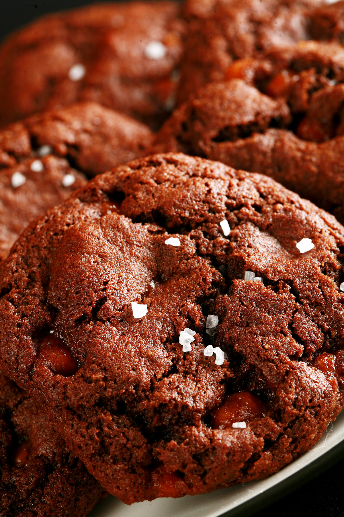 A small white plate, piled with Chewy, Gluten-Free Chocolate Cookies. Bits of caramel are visible within the cookies, and each is topped with a sprinkle of sea salt.