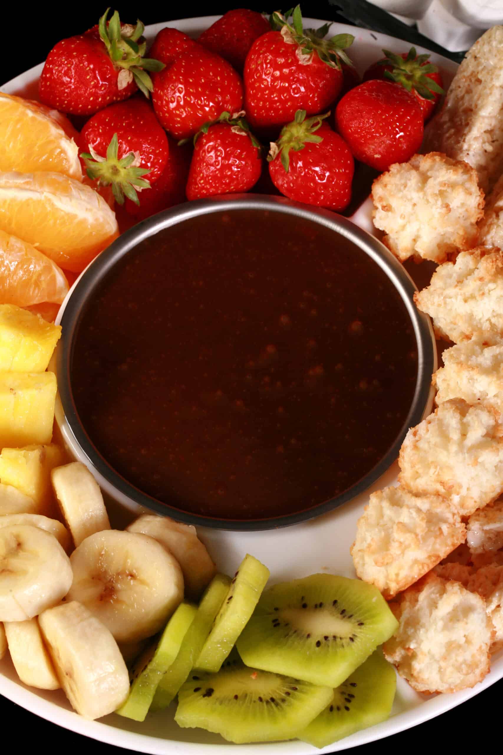 A bowl of easy chocolate fondue, surrounded by pineapple chunks, banana slices, kiwi slices, coconut macaroons, oranges, pretzels, marshmallows, and more.