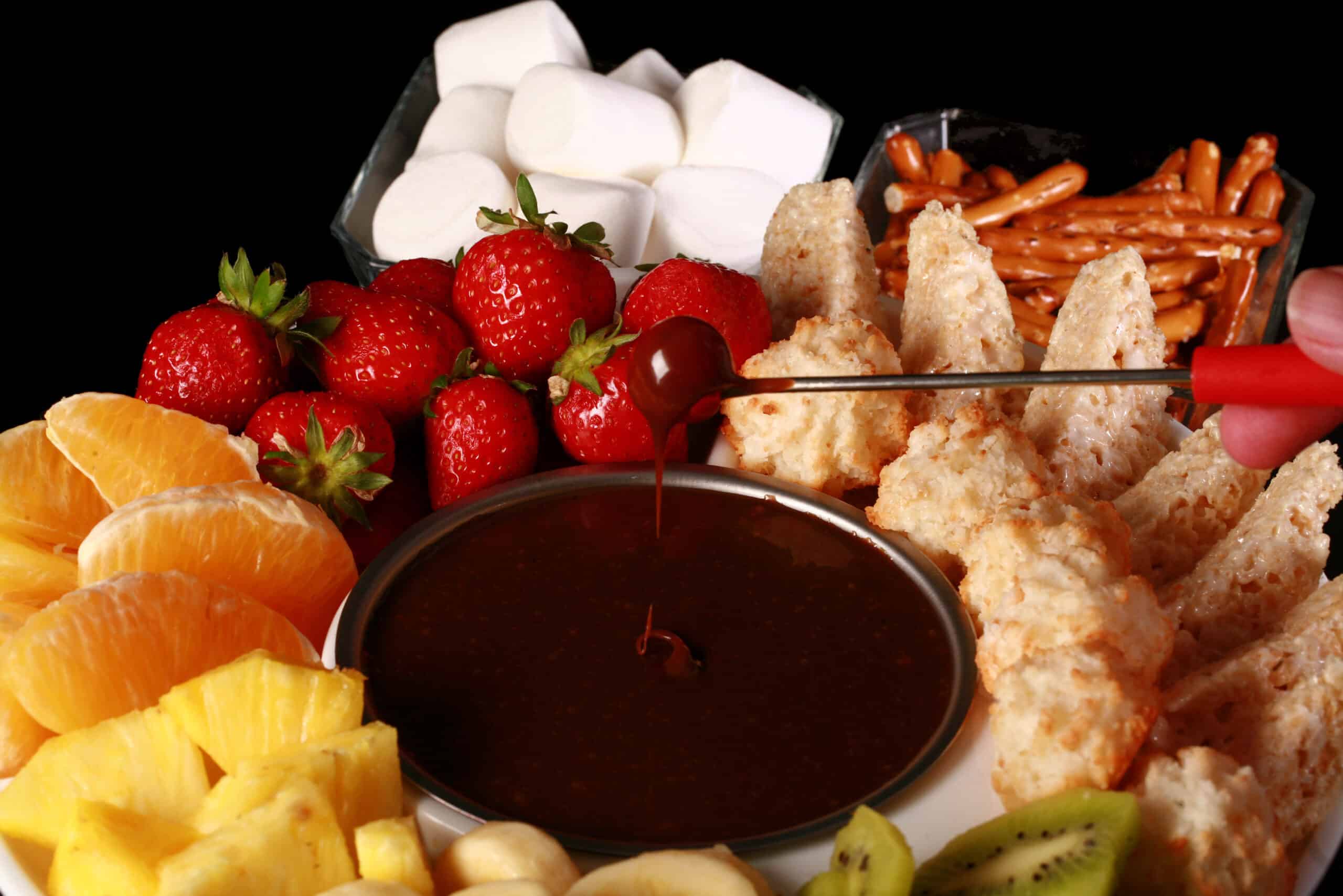 A strawberry being dipped into chocolate fondue, surrounded by pineapple chunks, banana slices, kiwi slices, coconut macaroons, oranges, pretzels, marshmallows, and more.
