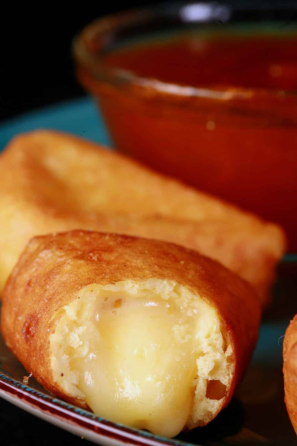 A plate of battered gluten free deep fried brie cheese wedges, around a bowl of apricot preserves. One has a bite, with brie oozing out.