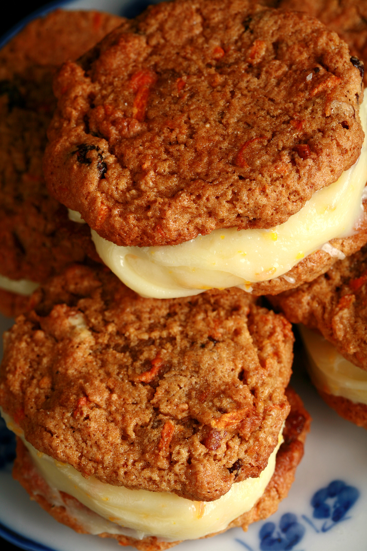 A small blue and white plate with a stack of carrot cake sandwich cookies on it. The filling is ivory coloured and has flecks of orange zest throughout