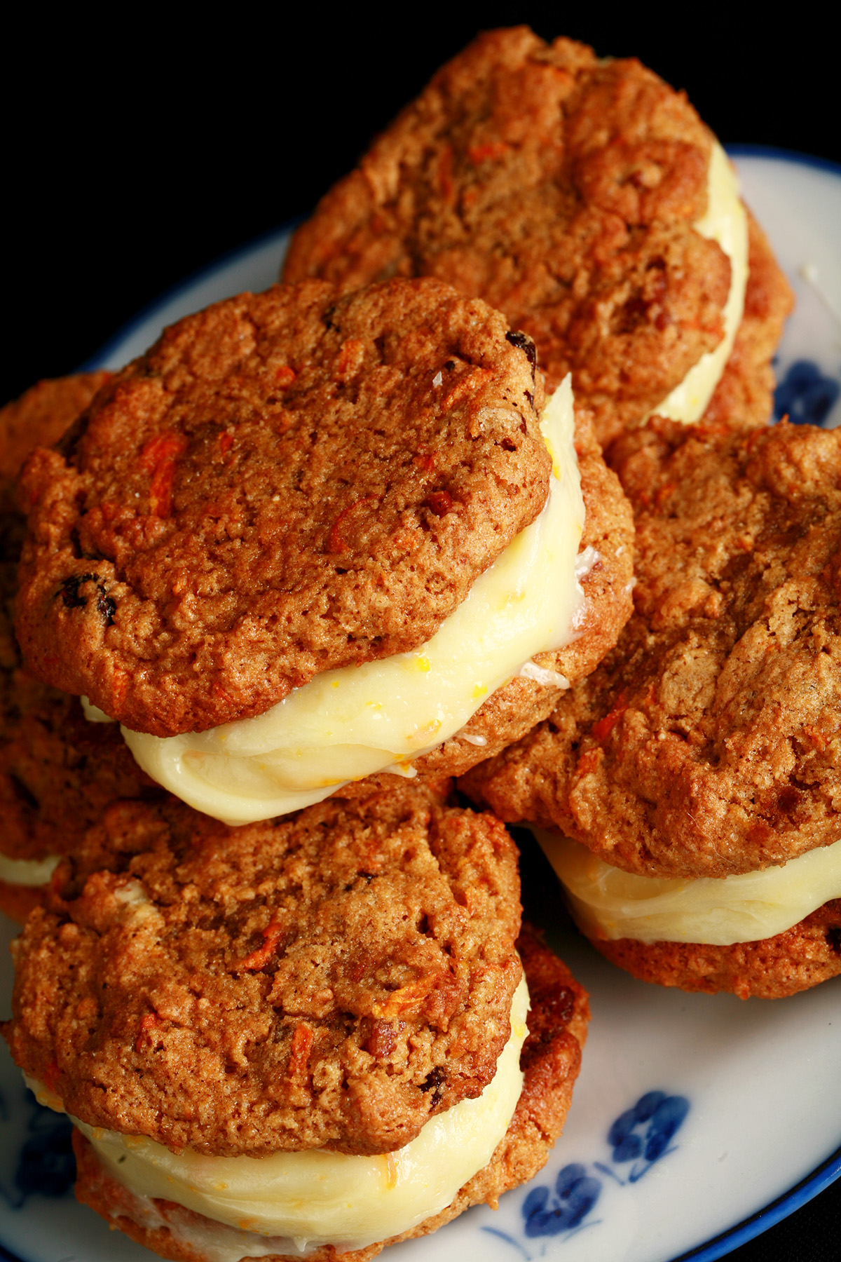 A small blue and white plate with a stack of carrot cake sandwich cookies on it. The filling is ivory coloured and has flecks of orange zest throughout.