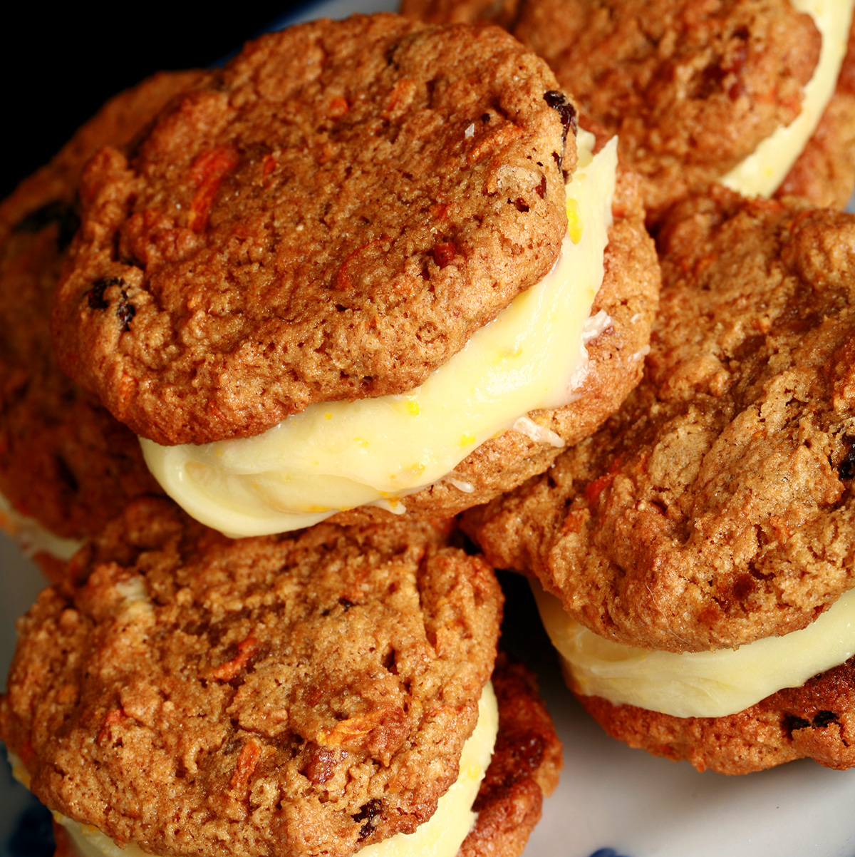 "A small blue and white plate with a stack of gluten-free carrot cake cookies on it. The filling is ivory coloured and has flecks of orange zest throughout.