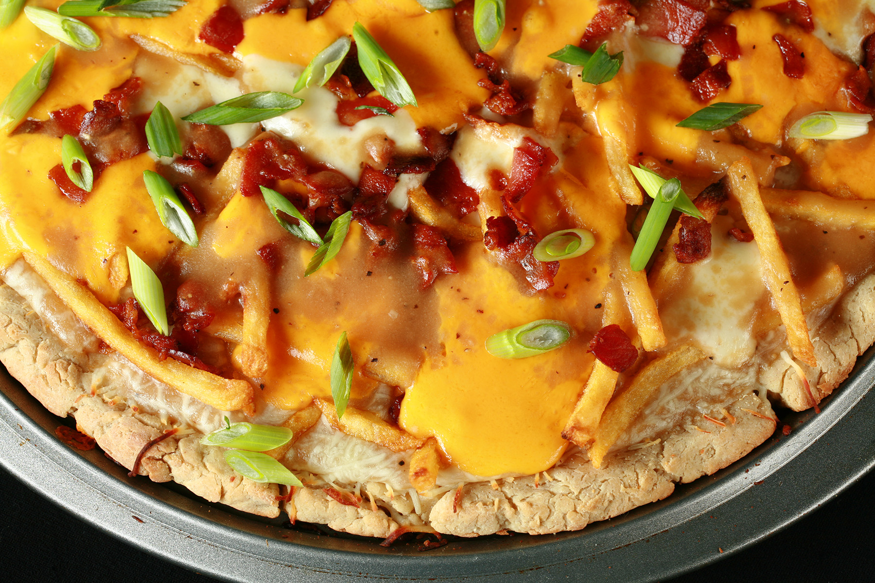 A close up view of a gluten-free poutine pizza - A pizza crust topped with gravy, fries, cheese curds, chredded cheese, bacon, and green onions!