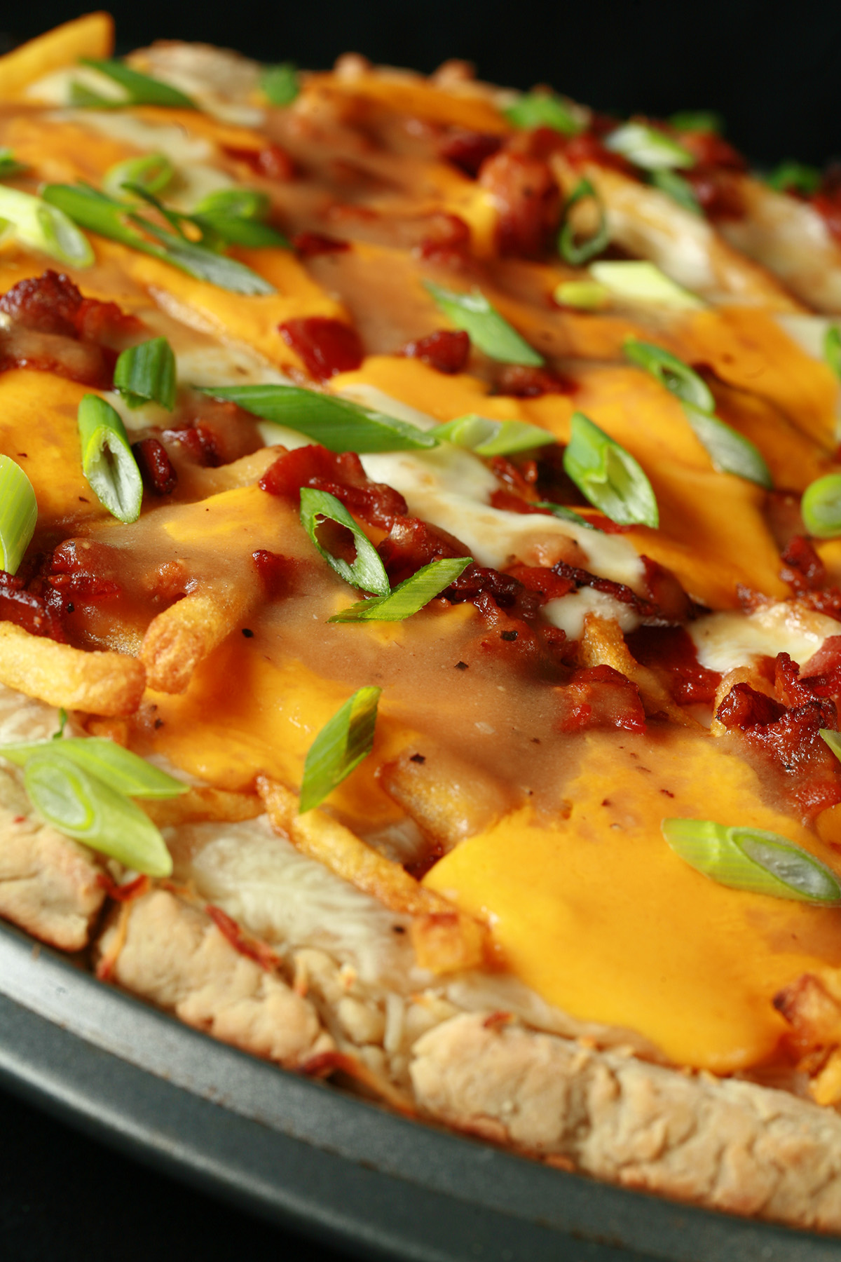 A close up view of a gluten-free bacon poutine pizza - A pizza crust topped with gravy, fries, cheese curds, chredded cheese, bacon, and green onions!