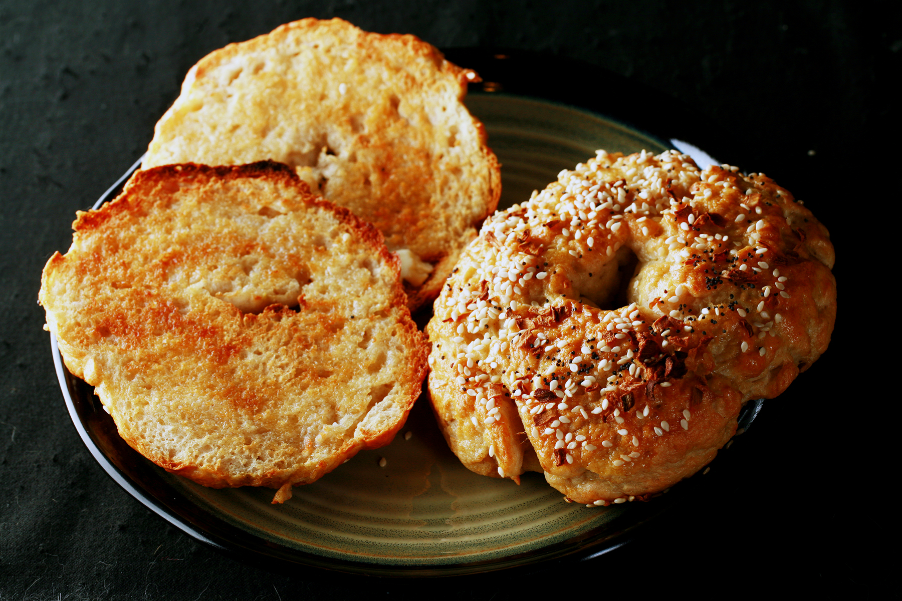 A close up view of two toasted gluten-free bagels on a plate. One is coated with everything seasoning.