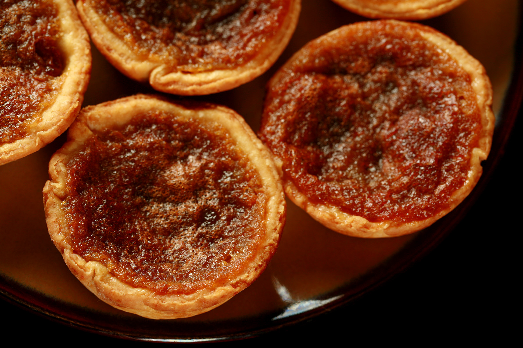 A brown plate is crowded with gluten-free butter tarts - mini tarts with a gooey sugar filling and raisins
