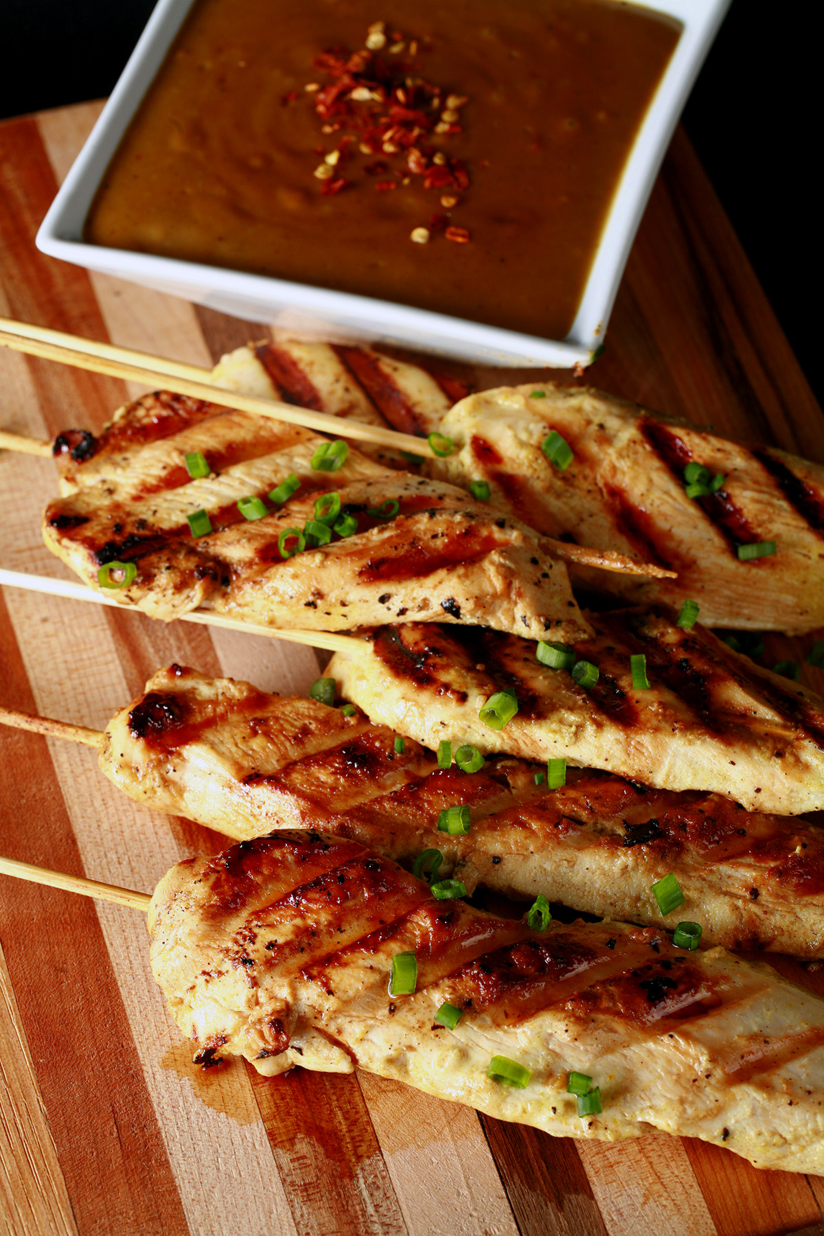 Several skewers of gluten-free chicken satay on a striped cutting board. They are sprinkled with green onions and sitting next to a bowl of peanut sauce.