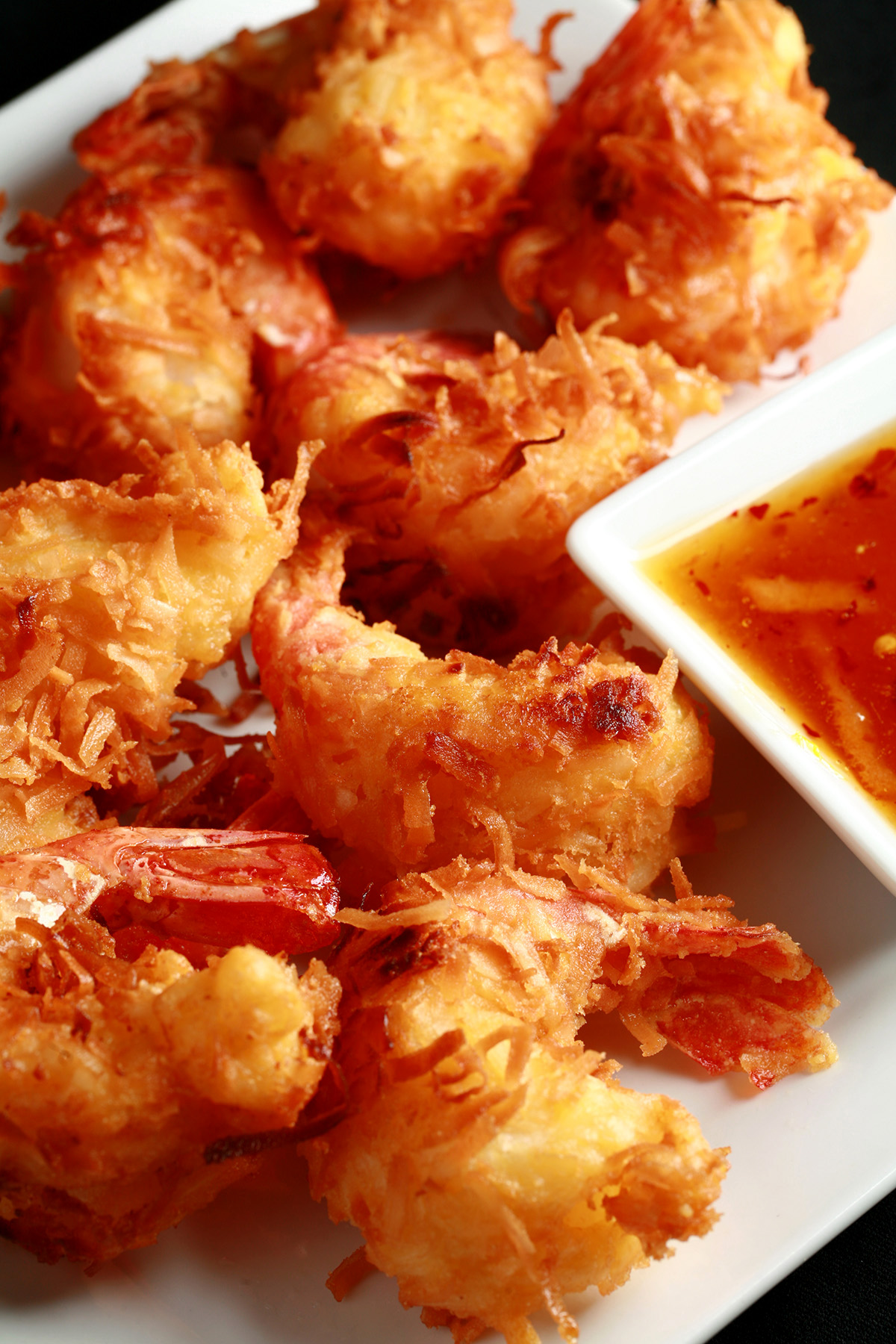 A white plate is covered with golden brown gluten-free coconut shrimp. There is a small bowl of spicy ginger-orange sauce on the same plate, as a dip.