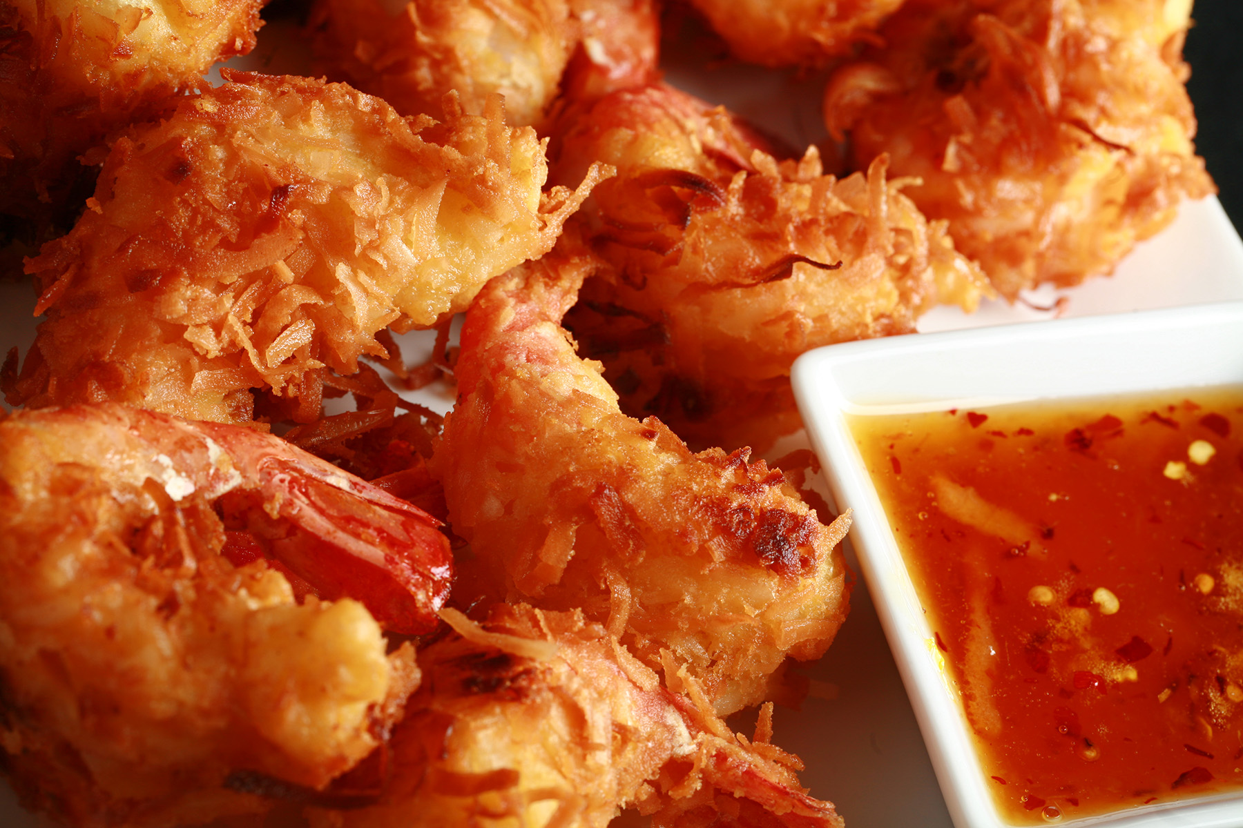 A white plate is covered with golden brown gluten-free coconut shrimp. There is a small bowl of spicy ginger-orange sauce on the same plate, as a dip.