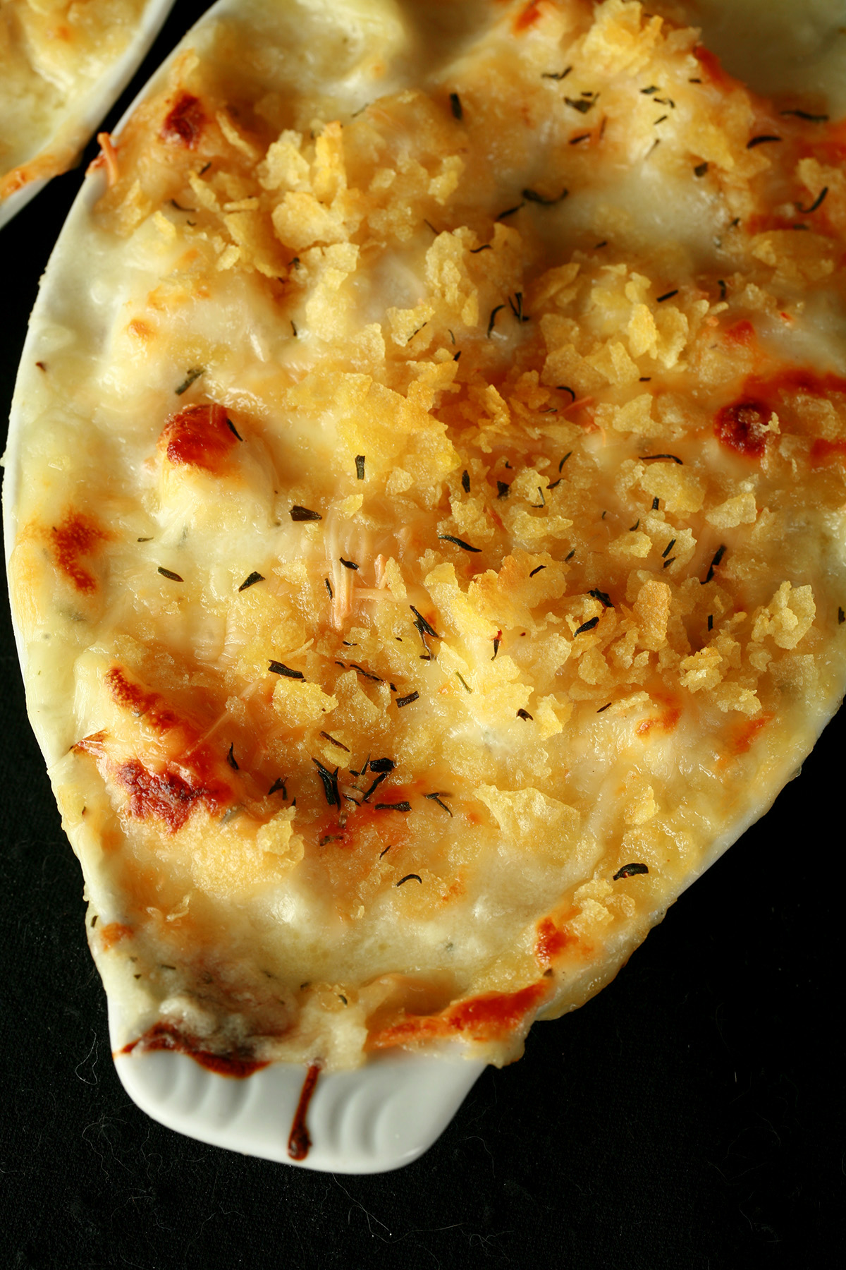 Close up view of a ceramic ramekin of gluten-free cod au gratin. Cod is in a creamy, cheesy sauce, topped with crushed chips, more cheese, and savoury.