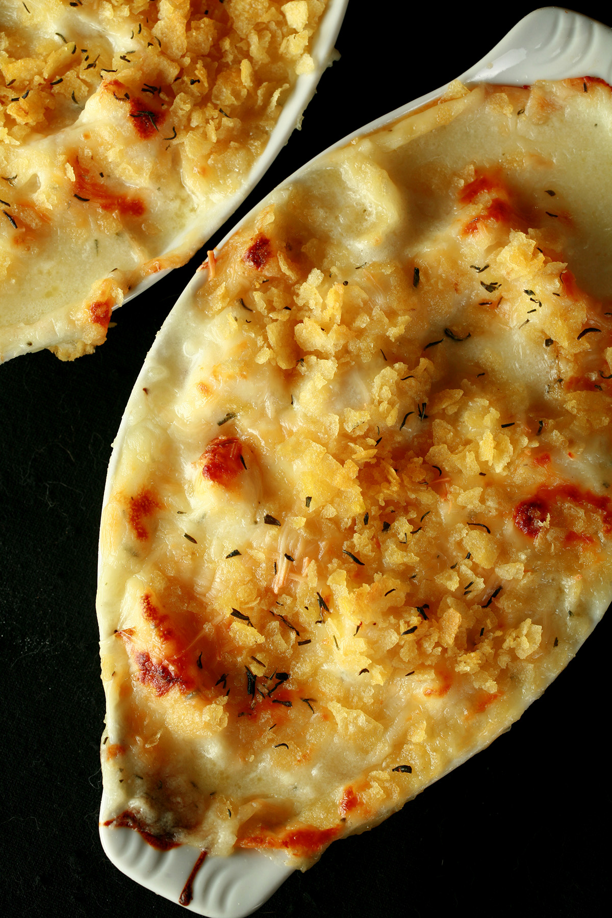 Two oblong ceramic ramekins of gluten-free cod au gratin. Cod is in a creamy, cheesy sauce, topped with crushed chips, more cheese, and savoury.