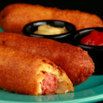 3 gluten-free corndogs on a green plate, with little bowls of ketchup and mustard.