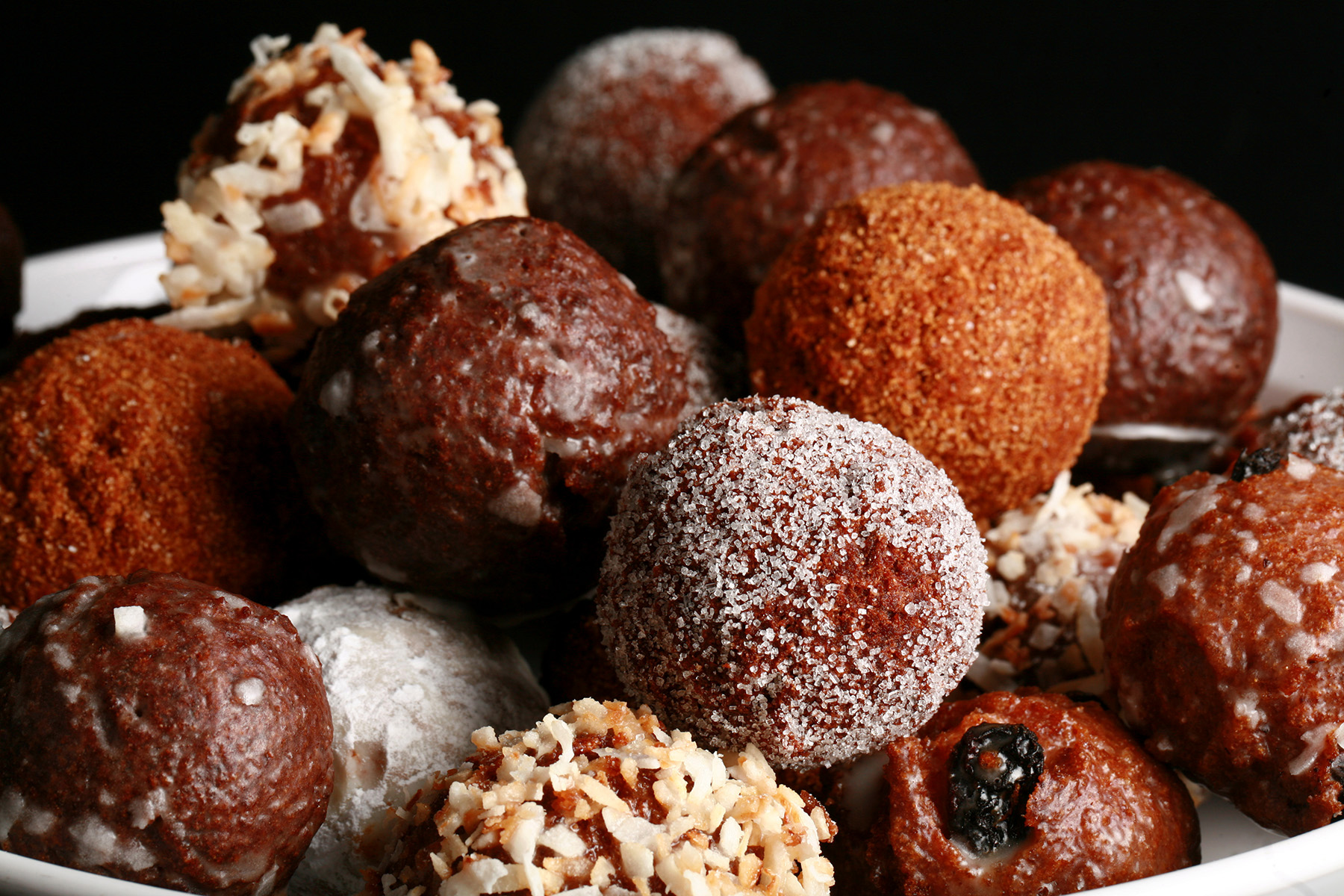 A close up view of an assortment of gluten-free doughnut holes: Toasted coconut, chocolate glazed, cinnamon sugar, powdered sugar, and dutchie versions are all visible.
