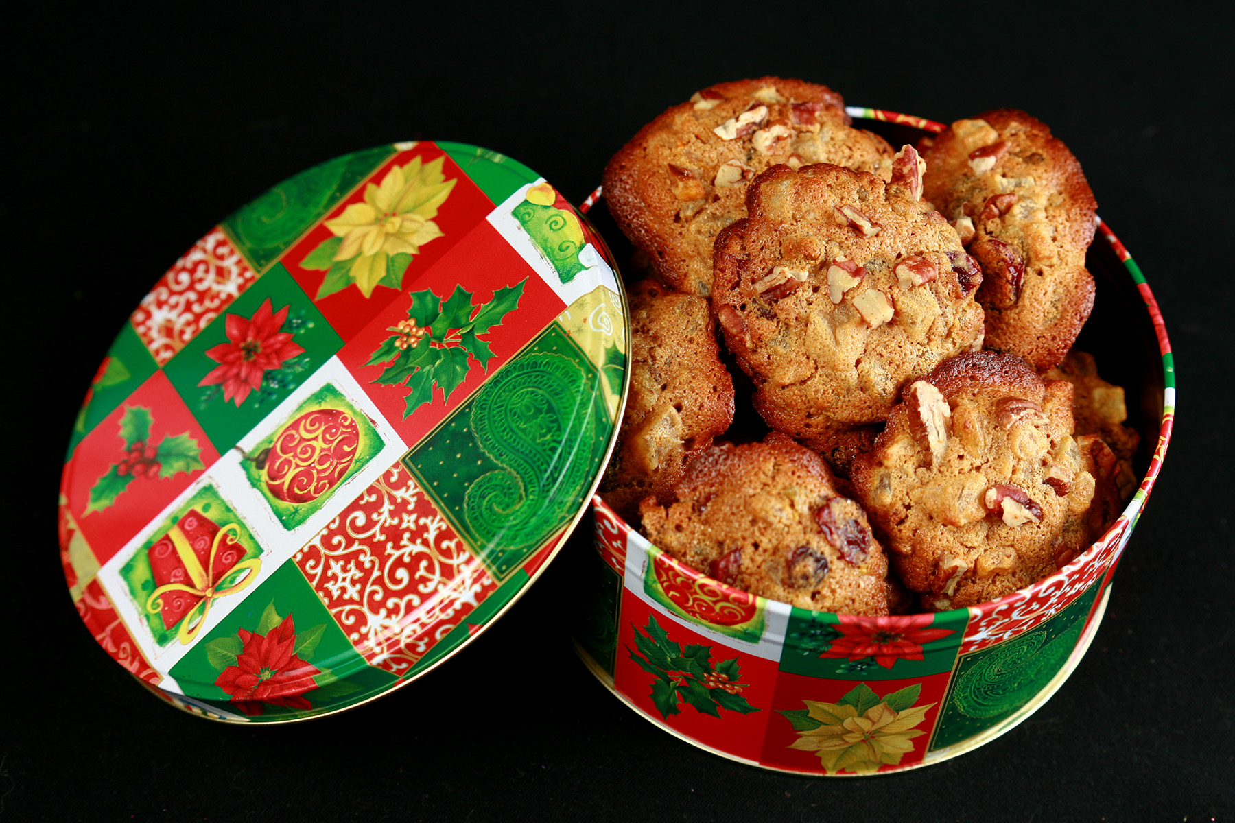 A festive green and red holiday container is full of cookies. The cookies have visible pieces of fruits and nuts throughout.