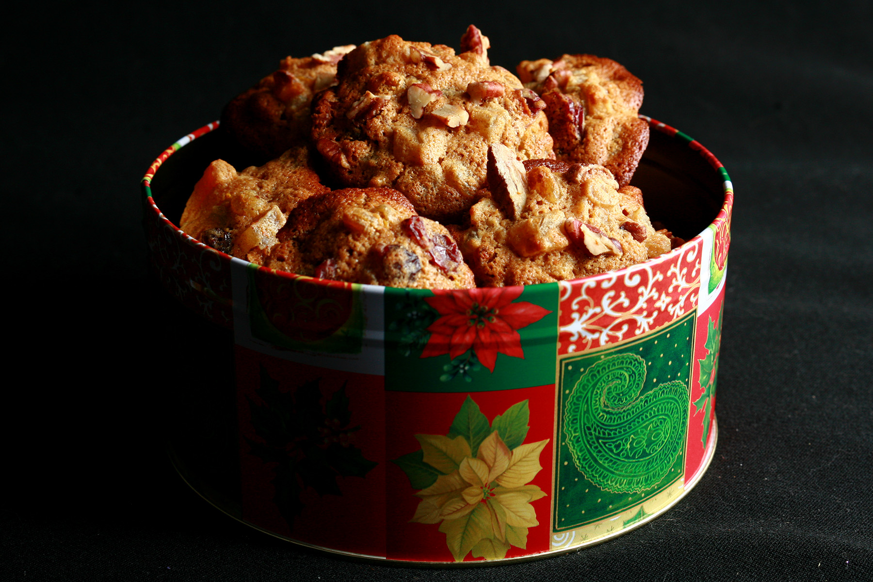 "A festive green and red holiday container is full of cookies. The cookies have visible pieces of fruits and nuts throughout.