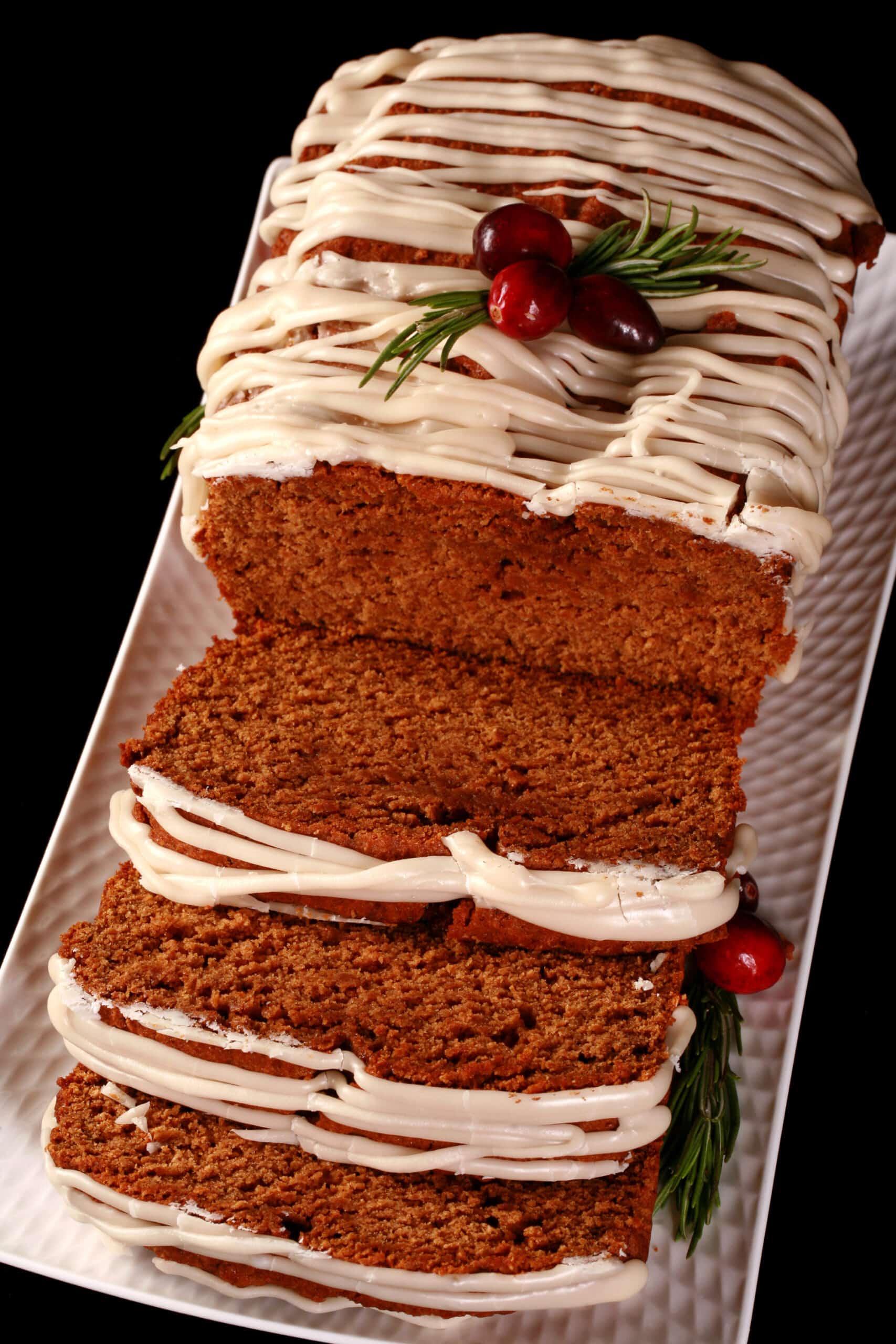 A sliced loaf of gluten-free gingerbread loaf, drizzled with vanilla glaze and garnished with rosemary and cranberries.