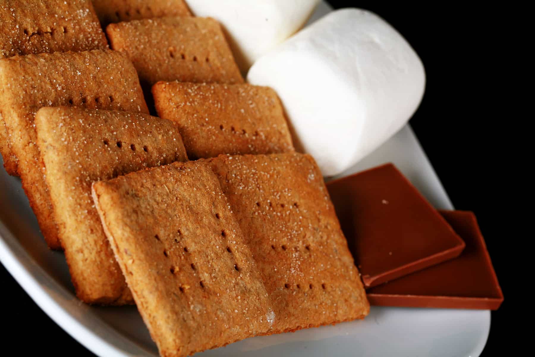 A white plate with rows of square shaped gluten-free graham crackers, a couple large marshmallows, and a piece of chocolate.