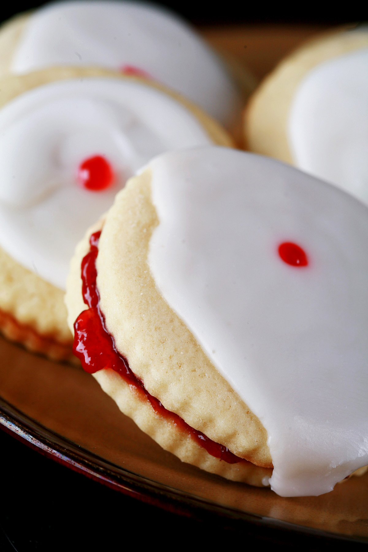 Close up view of a plate of gluten-free imperial cookies: Sandwich cookies filled with raspberry jam, frosted with a white glaze, and finished off with a dot of red gel in the center.