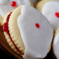 Close up view of a plate of Gluten-Free Empire Biscuits - sandwich cookies filled with raspberry jam, frosted with a white glaze, and finished off with a dot of red gel in the center.