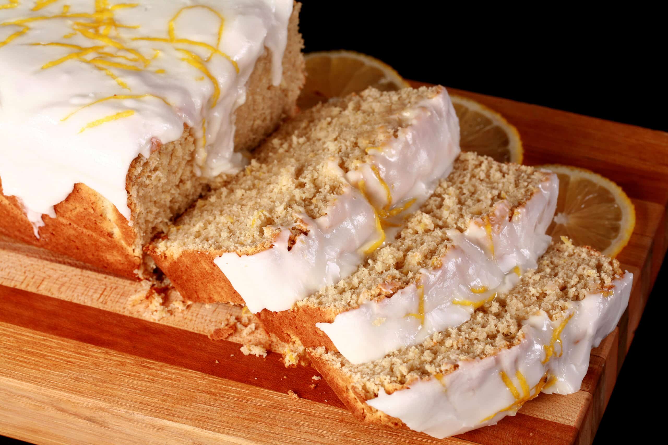 A sliced loaf of gluten-free lemon drizzle cake, with white glaze and lemon zest on top.