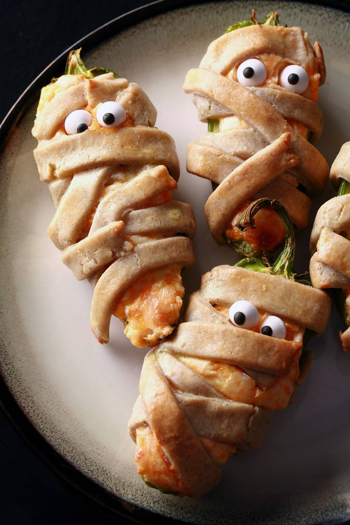 Several gluten free mummy jalapeno poppers on a plate. Each has 2 candy eyeballs.