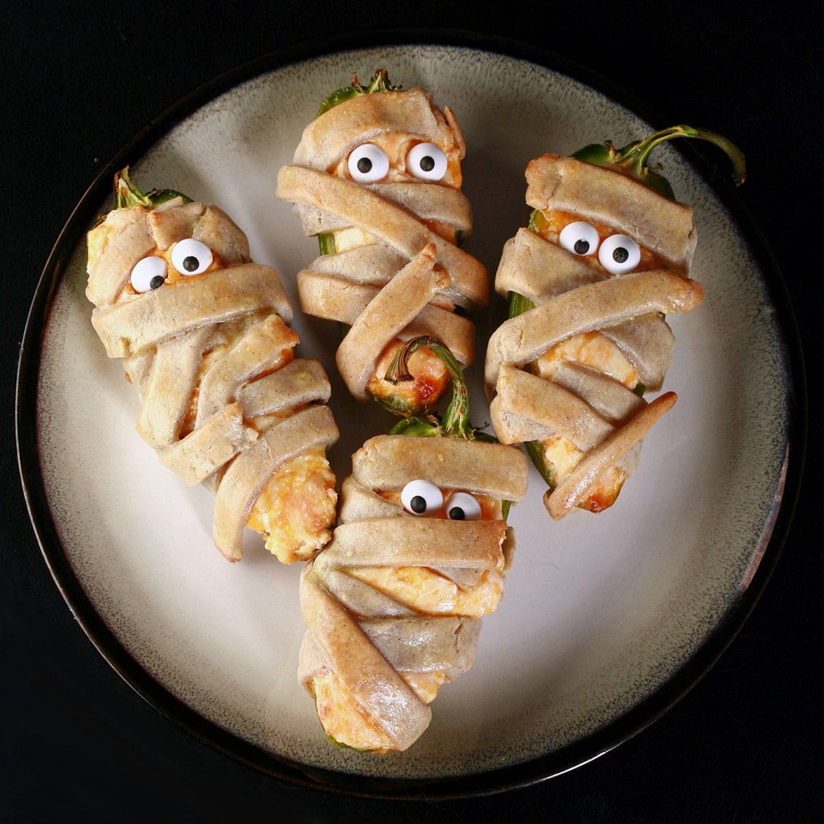 Several gluten free mummy jalapeno poppers on a plate. Each has 2 candy eyeballs.