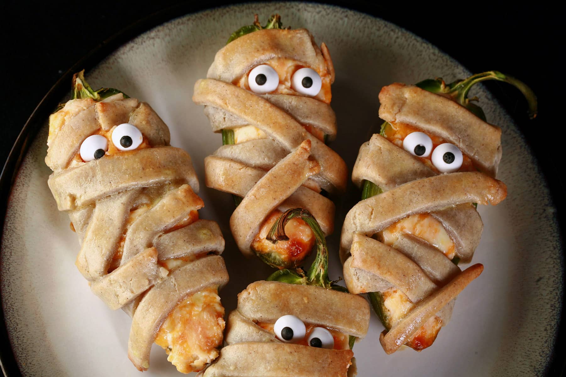 Several gluten free mummy jalapeno poppers on a plate. Each has 2 candy eyeballs.