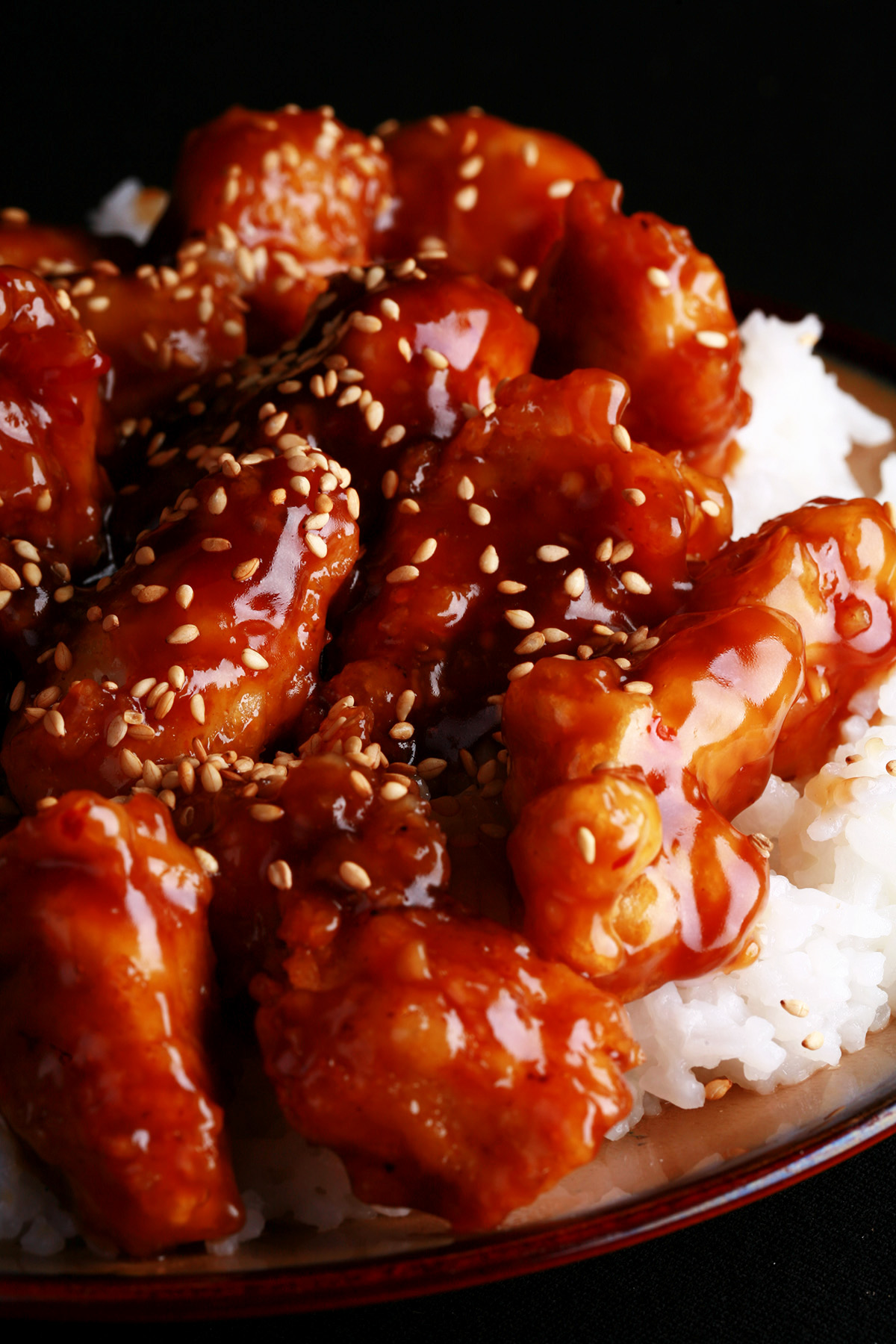 A close up view of a plate of white rice, topped with gluten-free sesame chicken. Chunks of battered and fried chicken in a glossy brown sauce, sprinkled with sesame seeds.