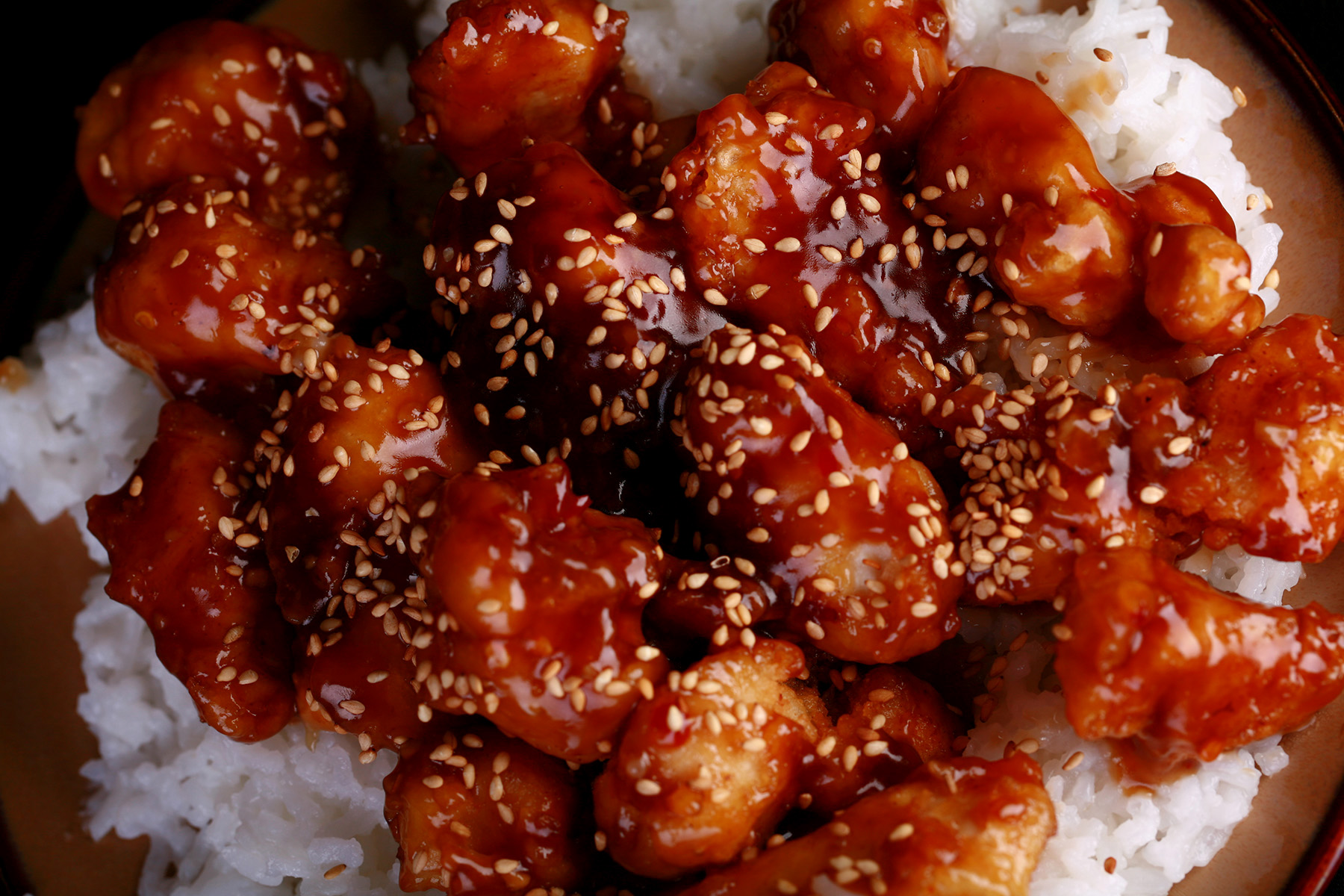 A close up view of a plate of white rice, topped with gluten-free sesame chicken. Chunks of battered and fried chicken in a glossy brown sauce, sprinkled with sesame seeds.