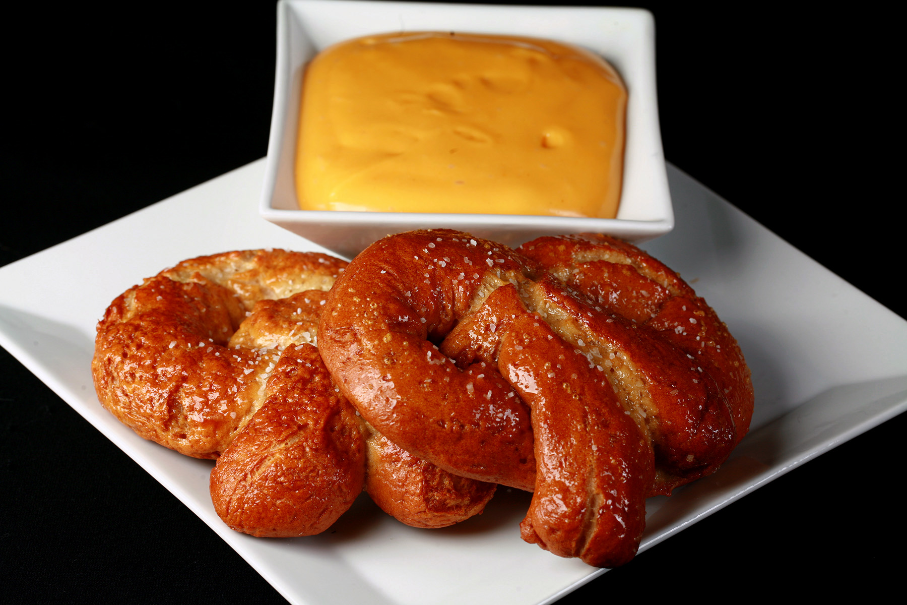 2 gluten-free soft pretzels on a plate, beside a bowl of mustard-cheddar dip.