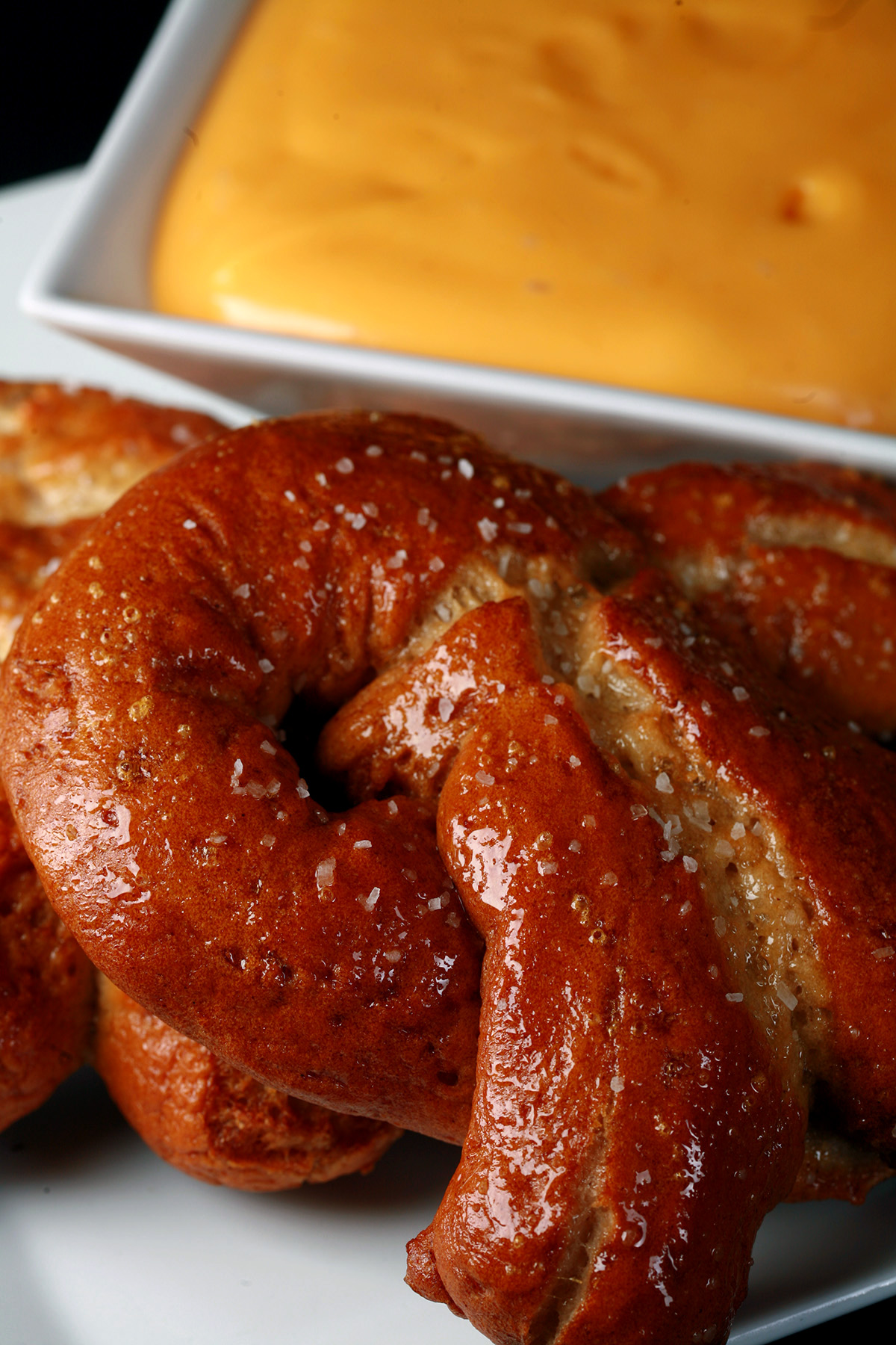 2 gluten-free soft pretzels on a plate, beside a bowl of mustard-cheddar dip.