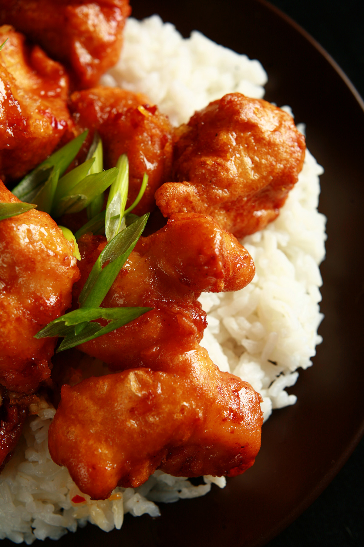 A plate of rice, covered with spicy orange chicken: Chunks of battered and deep fried chicken coated in a glossy orange sauce, garnished with sliced green onions.