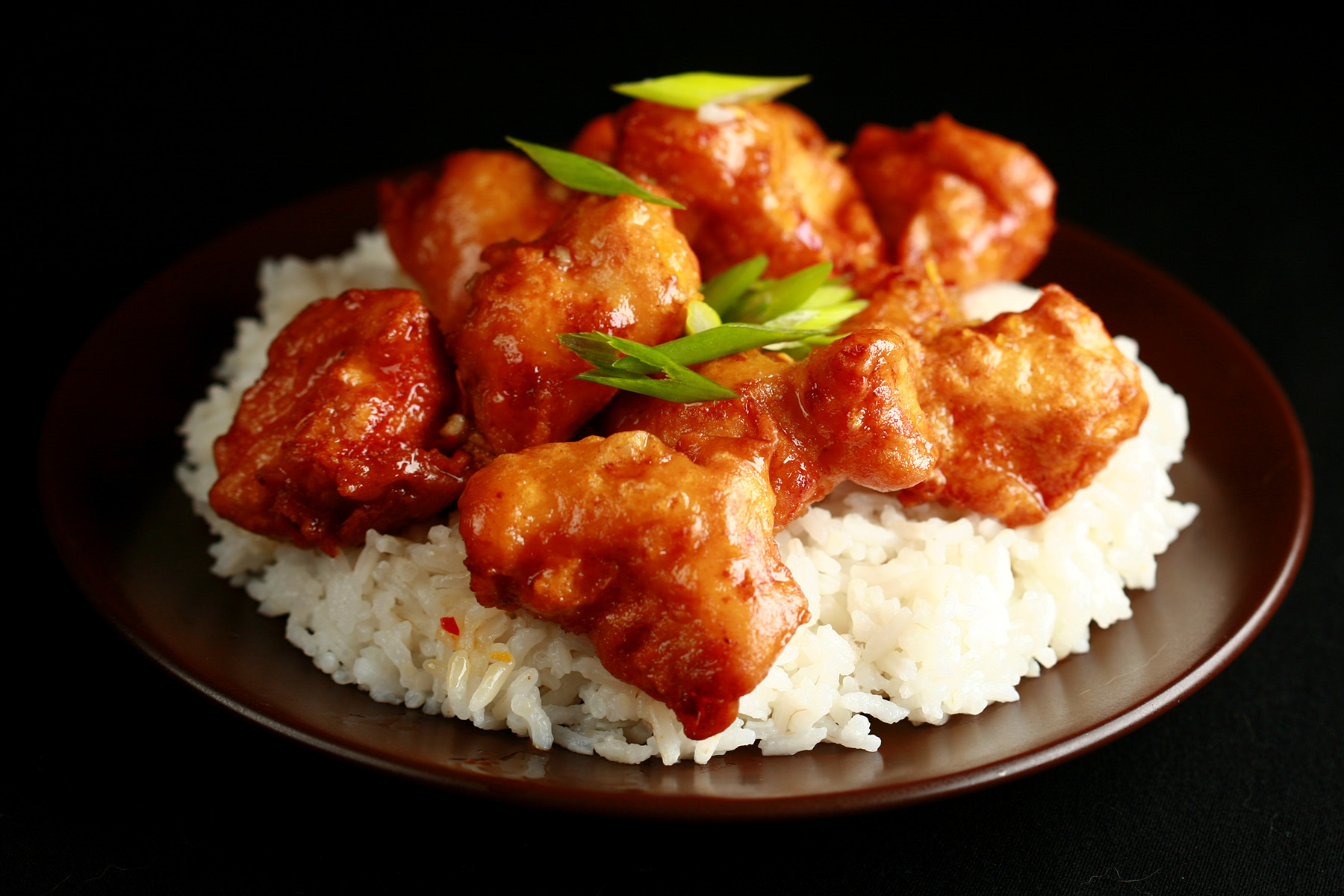 A plate of rice, covered with gluten-free spicy orange chicken: Chunks of battered and deep fried chicken coated in a glossy orange sauce, garnished with sliced green onions.