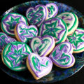 Close up view of a plate of heart shaped gluten-free sugar cookies. They are decorated with swirls of teal and lavender over a base of white frosting.