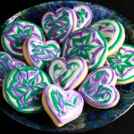 Close up view of a plate of heart shaped gluten-free sugar cookies. They are decorated with swirls of teal and lavender over a base of white frosting.