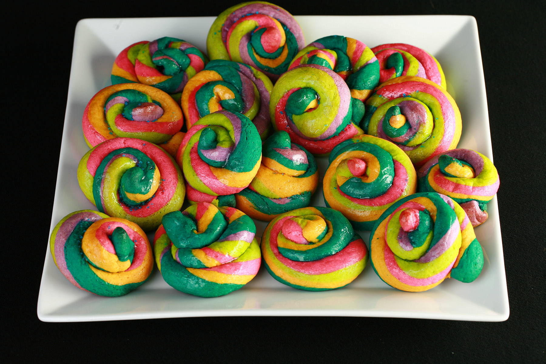 A white, square plate piled with gluten-free unicorn poop cookies. They are brightly coloured cookie "poops" made from brightly coloured twists of cookie dough.
