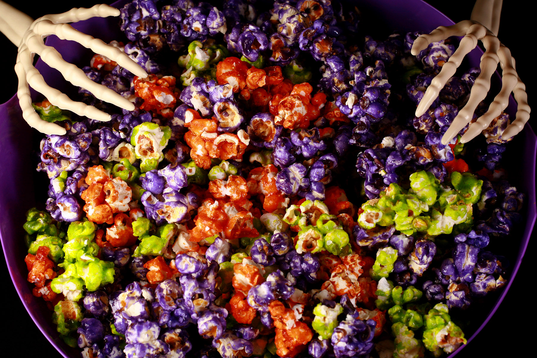 A bowl of Halloween glazed popcorn - orange, purple, and lime green mixed together. A pair of skeleton hand serving spoons are visible.