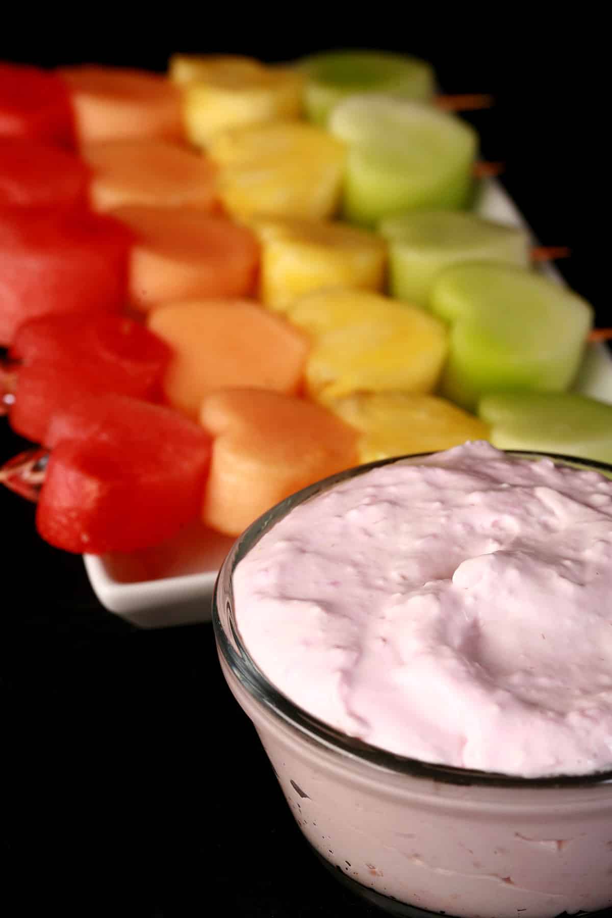 Several skewers of heart shaped fruit - watermelon, cantaloupe, pineapple, and honeydew - next to a bowl of raspberry cheesecake dip.