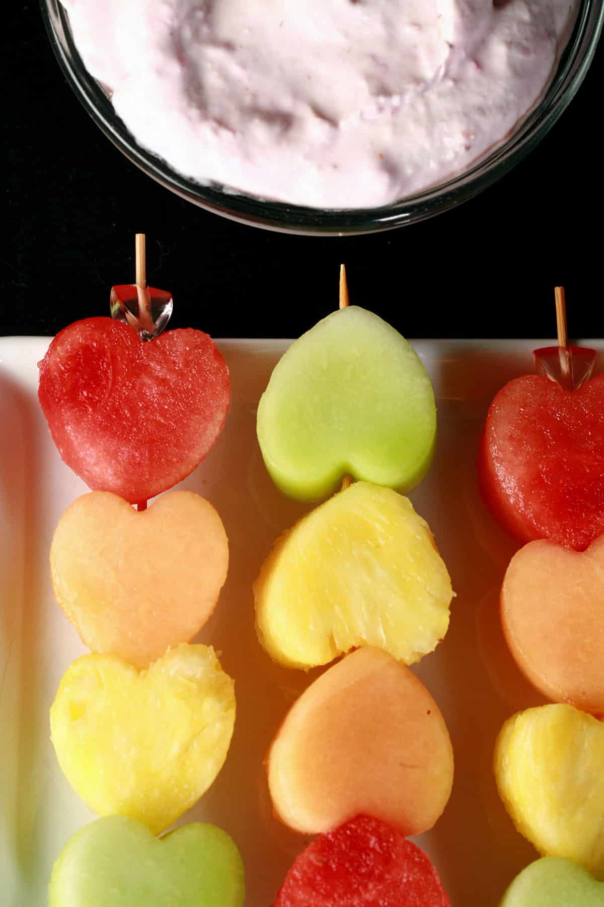 Several skewers of heart shaped fruit - watermelon, cantaloupe, pineapple, and honeydew - next to a bowl of raspberry cheesecake dip.