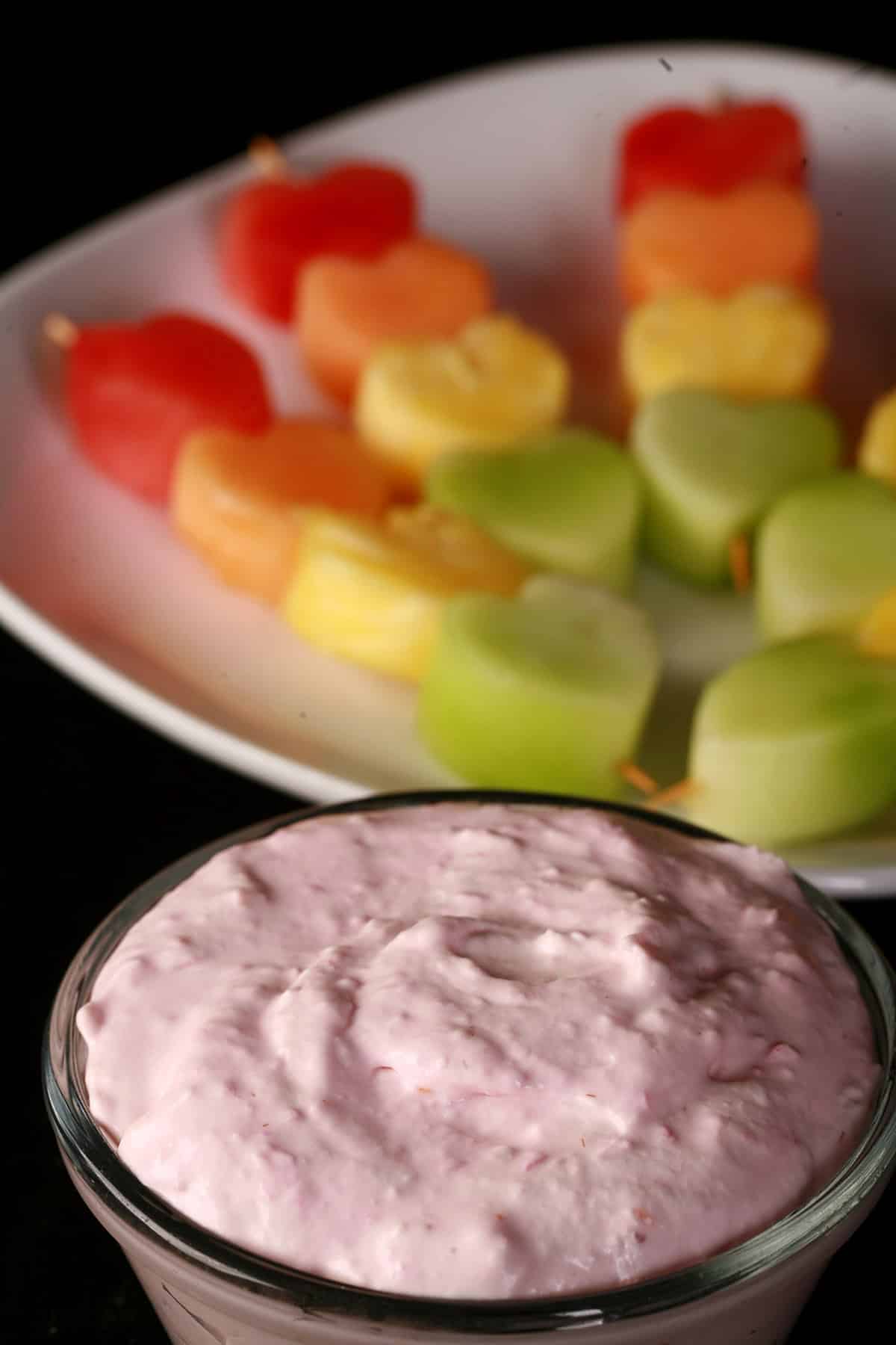 Several heart shaped fruit skewers- watermelon, cantaloupe, pineapple, and honeydew - next to a bowl of raspberry cheesecake dip.