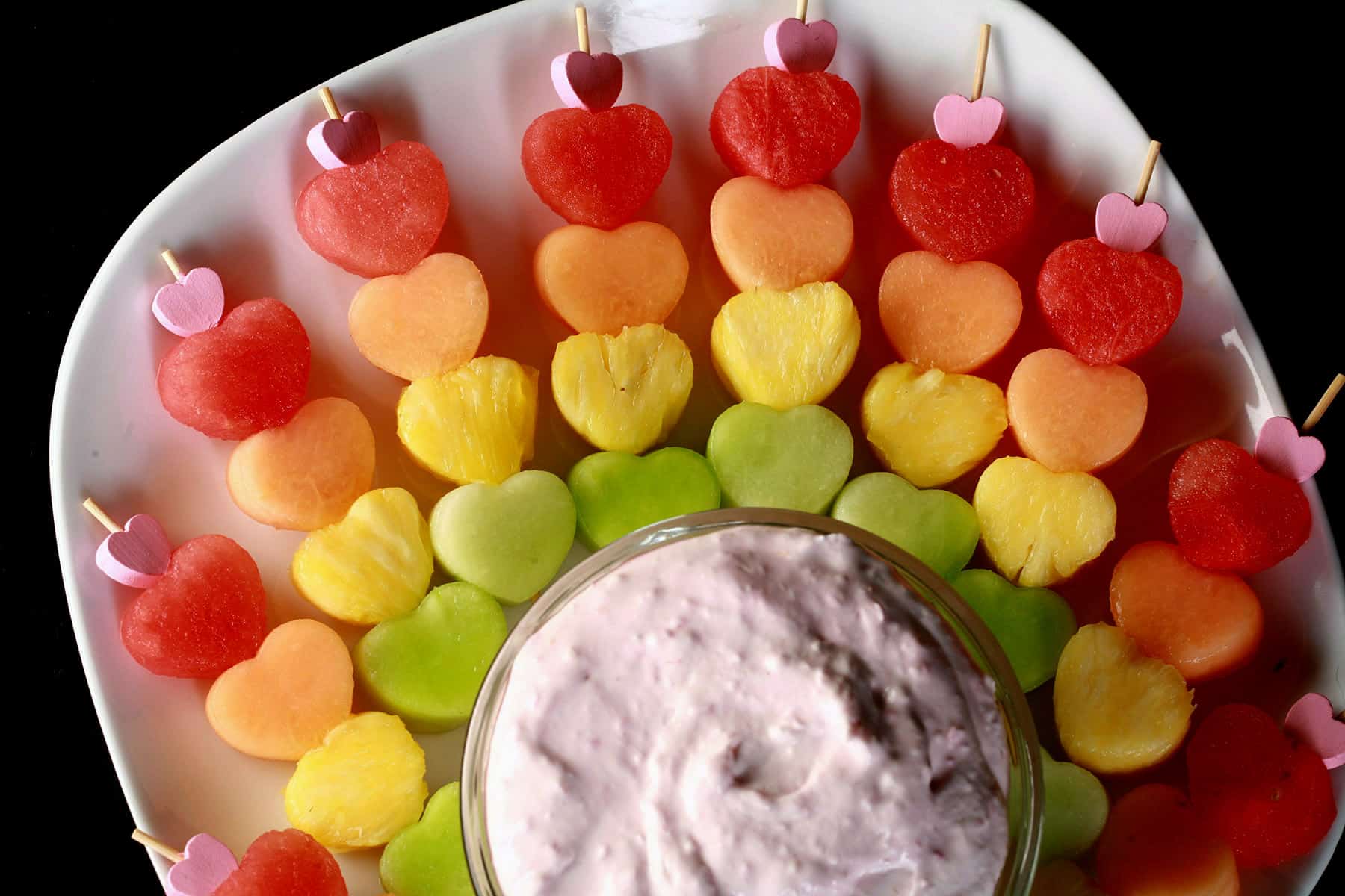 Several skewers of heart shaped fruit - watermelon, cantaloupe, pineapple, and honeydew - next to a bowl of raspberry cheesecake dip.