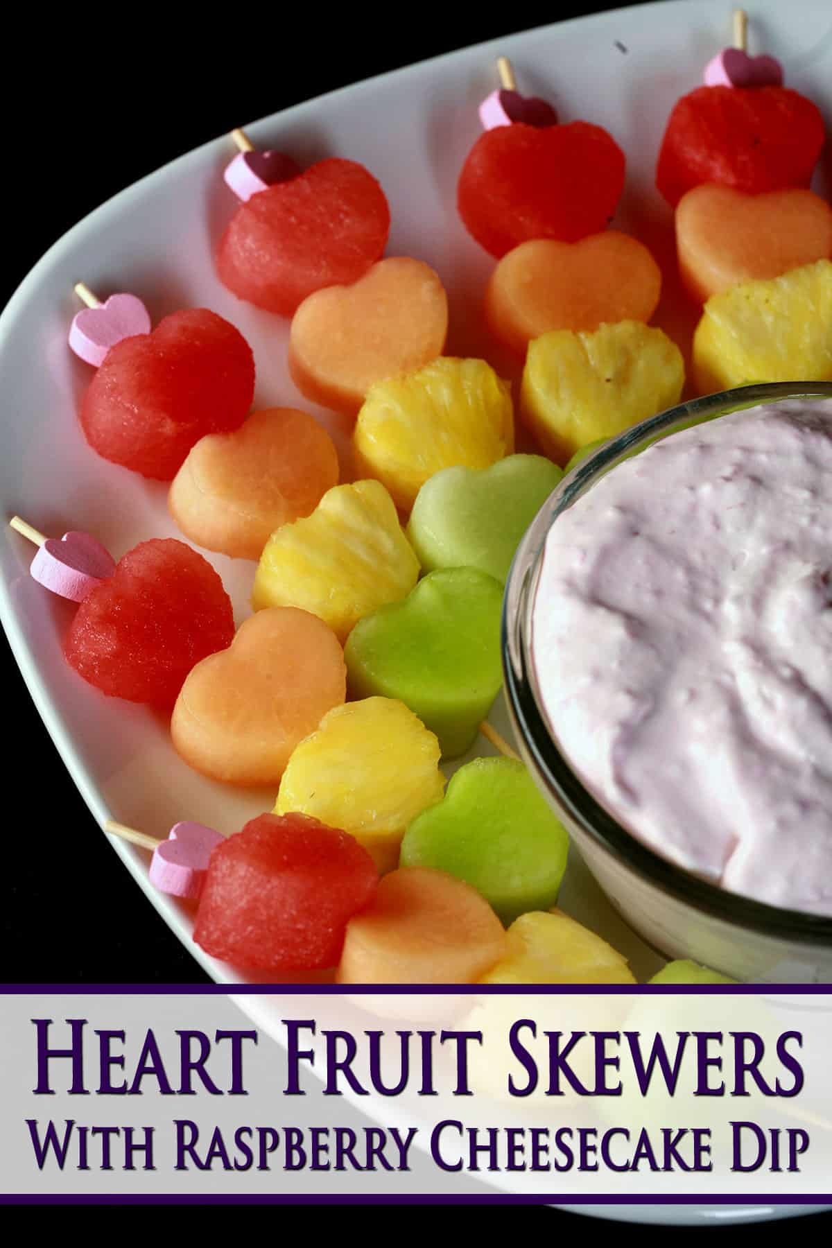 Several heart shaped fruit skewers- watermelon, cantaloupe, pineapple, and honeydew - next to a bowl of raspberry cheesecake dip.