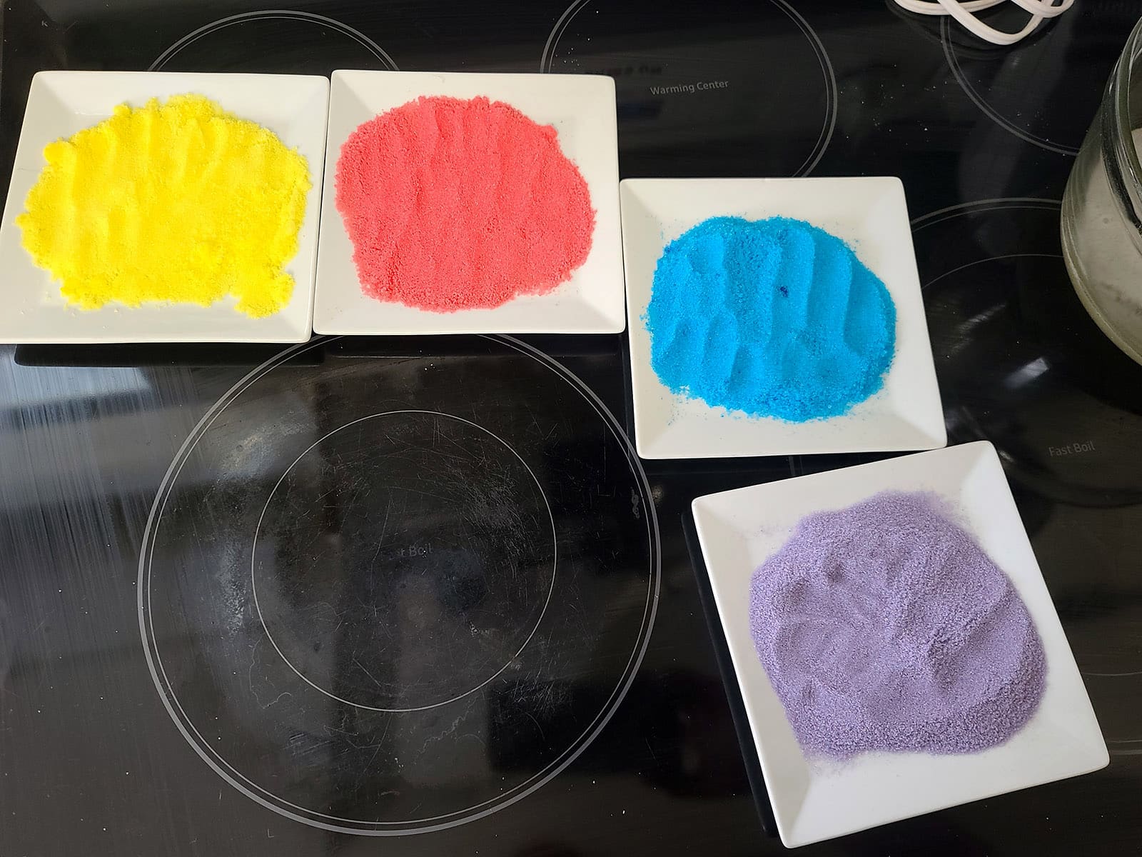 4 little plates, each with a different color of sugar on it - pink, yellow, blue, and purple.