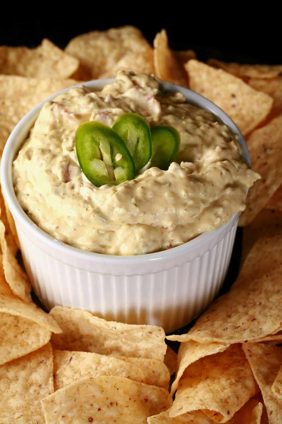 A ramekin of gluten free jalapeno dip, surrounded by corn chips and topped with sliced jalapenos.